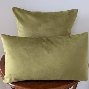 May include: Two olive green rectangular velvet cushions are stacked on a wooden chair. The cushions have a soft, plush appearance and a slight sheen. The chair has a dark wood finish and a curved design.