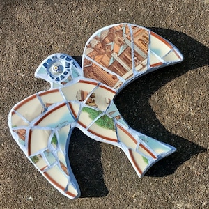 May include: A mosaic bird sculpture made from broken ceramic pieces. The bird features a mix of colors, including white, blue, brown, and green. The pieces are arranged to depict a bird in flight, with a detailed eye and wing design. The sculpture is set against a textured, gray surface.