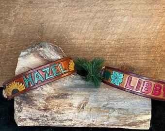 Puede incluir: Dos collares de perro de cuero personalizados con diseños florales. El collar superior es marrón con girasoles y el nombre "HAZEL" en letras verde azuladas. El collar inferior es marrón con flores rosas y el nombre "LIBBY" en letras rojas.