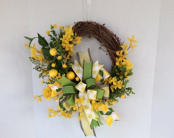 May include: A spring-themed wreath with yellow flowers, green leaves, and a yellow and white checkered bow with a green ribbon. The wreath is made of grapevine and has a lemon print ribbon.
