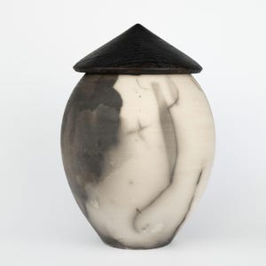 May include: A white ceramic urn with a black wooden lid. The urn has a unique, abstract design with gray and white swirls.