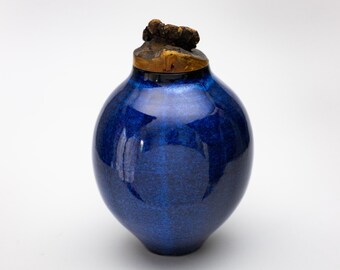 Könnte beinhalten: Eine tiefblaue Keramikvase mit einem Holzdeckel. Die Vase hat eine glatte, glänzende Oberfläche und eine abgerundete Form.