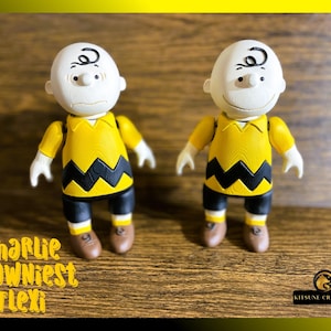 May include: Two yellow and black Charlie Brown figurines with white heads and black eyes. The figurines are standing on a wooden surface. The text "Charlie Brownniest Flexi" is visible in the lower left corner of the image.