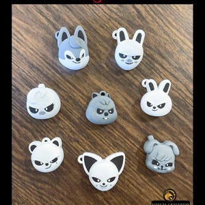 Puede incluir: Una colección de amuletos de personajes grises y blancos con expresiones enfadadas, incluyendo un lobo, conejos y un oso. La palabra "Stray Kids" está en la parte superior y "dominate" en la inferior en fuente gótica roja.