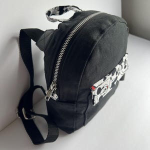 May include: Black mini backpack with a silver zipper and adjustable black straps. The backpack features a small handle with a black and white pattern. A white patch with a Mickey Mouse design is on the side.