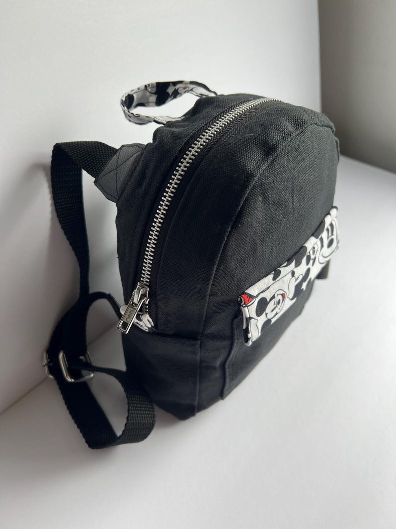 May include: Black mini backpack with a silver zipper and adjustable black straps. The backpack features a small handle with a black and white pattern. A white patch with a Mickey Mouse design is on the side.