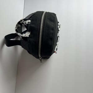 May include: Black fanny pack with a silver zipper and a black strap. The pack features a black and white patterned design on the sides and strap. The fanny pack is made of a durable material.