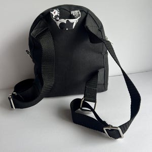 May include: A small, black backpack with adjustable straps and silver-colored metal hardware. The interior lining visible at the top of the bag features a white and black floral pattern. The bag has a zippered closure and is designed for carrying small items.