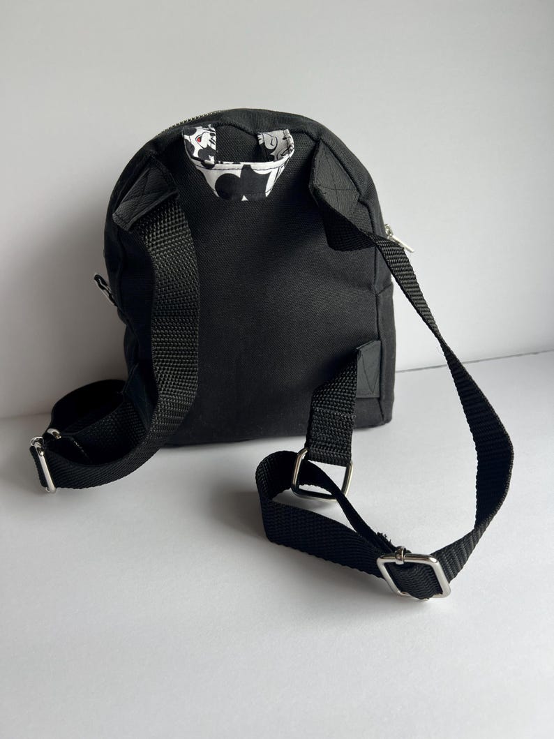 May include: A small, black backpack with adjustable straps and silver-colored metal hardware. The interior lining visible at the top of the bag features a white and black floral pattern. The bag has a zippered closure and is designed for carrying small items.