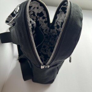 May include: Black canvas sling bag with a silver zipper and a black and white patterned interior. The bag has a black adjustable strap and a small pocket. The bag is open, revealing the interior design.
