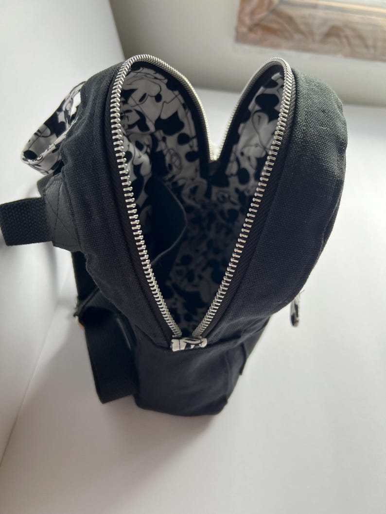 May include: Black canvas sling bag with a silver zipper and a black and white patterned interior. The bag has a black adjustable strap and a small pocket. The bag is open, revealing the interior design.