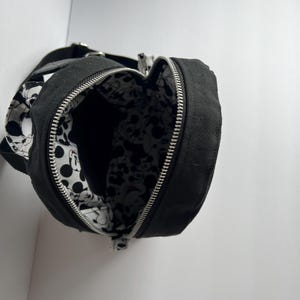 May include: Black canvas crossbody bag with a silver zipper and a black strap. The interior lining features a black and white Mickey Mouse pattern. The bag is open, revealing the interior design. The bag is designed to be worn over the shoulder.