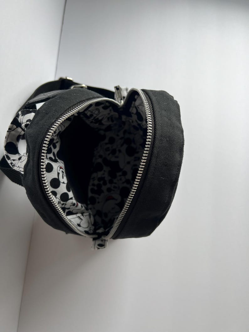 May include: Black canvas crossbody bag with a silver zipper and a black strap. The interior lining features a black and white Mickey Mouse pattern. The bag is open, revealing the interior design. The bag is designed to be worn over the shoulder.