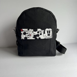 May include: A small black backpack with a rectangular front pocket featuring a Mickey Mouse pattern in black, white, and red. The backpack has a top handle, adjustable straps, and a zipper closure.