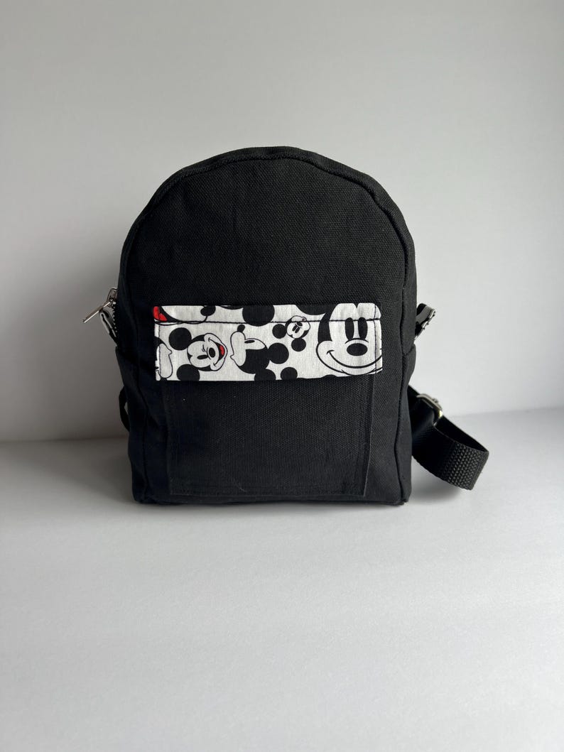 May include: A small black backpack with a rectangular front pocket featuring a Mickey Mouse pattern in black, white, and red. The backpack has a top handle, adjustable straps, and a zipper closure.