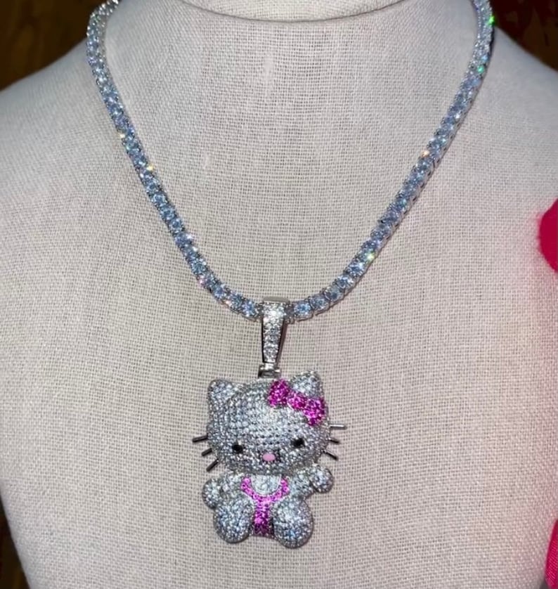 May include: A silver chain necklace with a white and pink Hello Kitty pendant. The pendant is covered in small clear stones and has a pink bow.