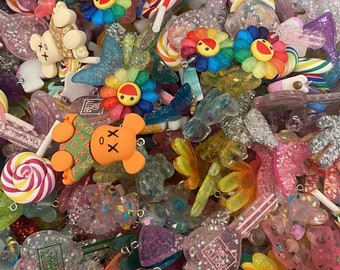 May include: A colorful assortment of resin charms, including rainbow flower faces, teddy bears, lollipops, and butterflies. The charms are in various colors, including orange, pink, yellow, and blue. Some charms have glitter accents and the text "PRETTY GIRL".