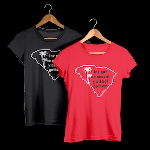 May include: Two t-shirts featuring a white outline of the state of South Carolina with palm trees and the text "I ne got no accent y'all boi gottem". The shirt on the left is black and the shirt on the right is red.