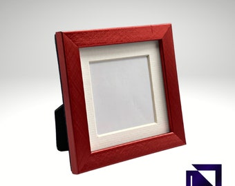 May include: A square red picture frame with a white mat and a black easel back. The frame is angled slightly, showcasing its textured surface. The logo "FRAMES & MORE" is in the bottom right corner.