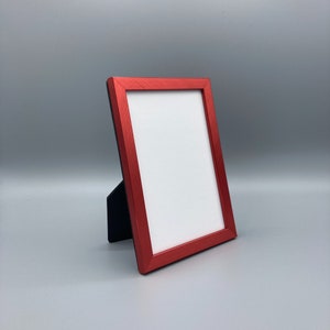 May include: A red picture frame with a black stand. The frame is made of metal and has a smooth, glossy finish.