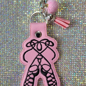 Puede incluir: Llavero de cuero rosa con un diseño bordado en negro de zapatos de ballet. El llavero tiene un anillo de metal plateado y un borla de peluche rosa.