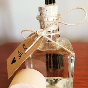 Puede incluir: Una botella de vidrio transparente con un tapón de corcho y una etiqueta de papel marrón que dice "Made with love". La botella está atada con un cordel y tiene un palo difusor de madera.