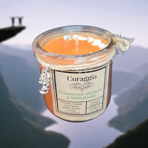 Puede incluir: Una vela de vidrio en un frasco con una etiqueta marrón que dice "Coraggio Frankincense and Myrrh & White Howlite". La etiqueta también indica que el aroma es "Scentual Relaxation" y que el peso neto es de "0,4 oz (11 g)". La vela está decorada con un amuleto plateado en forma de instrumento musical.