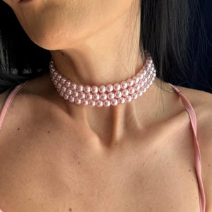 Può includere: Una collana girocollo a tre fili composta da perle artificiali rotonde di colore rosa chiaro. La collana aderisce al collo, mostrando una finitura uniforme e lucida su ogni singola perla. Il design è formato da tre file parallele.