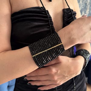 May include: A black beaded choker, a long beaded necklace with a teardrop pendant, and two wide beaded cuff bracelets. Each piece is constructed from small, faceted black beads and features a thin, horizontal gold-toned metal bar accent.