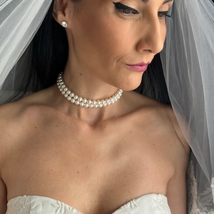 Può includere: Un abito bianco senza spalline con intricati ricami floreali, una collana girocollo di perle a doppio filo e un velo bianco trasparente con un ampio bordo in pizzo. Il velo ricade sulle spalle, incorniciando lo scollo e la collana di perle.