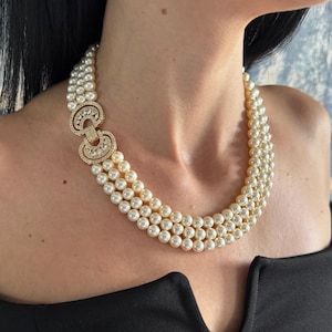 May include: A three-strand pearl necklace featuring cream-coloured round pearls and a gold-toned metal clasp on the side. The clasp is circular, embellished with small clear stones, and connects the three strands of pearls in a layered arrangement.
