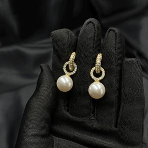 Puede incluir: Un par de pendientes colgantes con una perla blanca redonda suspendida de un aro circular de tono dorado. El aro está incrustado con pequeñas piedras claras y brillantes. El diseño muestra una combinación clásica de perlas y detalles metálicos.