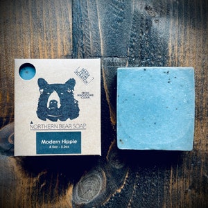 May include: A bar of blue soap with black flecks, labeled "Modern Hippie" and "4.5oz - 5.0oz". The soap is packaged in a brown cardboard box with a black bear illustration and the text "Made from Scratch, Fresh, Wholesome, Clean, Northern Bear Soap".