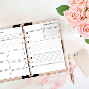 May include: A pink and white planner with a gold binder ring. The planner is open to a page titled "In Depth Bible Study" with sections for book, chapter, background passage, context, interpretation, additional notes, prayer, thoughts and feelings.