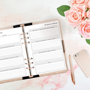 May include: An open planner with a beige cover and gold rings. The pages are filled with a printable scripture analysis worksheet. The worksheet includes prompts for reflection and journaling. The planner is on a white surface with a pink rose bouquet and a gold phone.