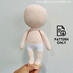 May include: A crocheted doll body in a light peach color, with white shorts. The doll is standing on a small wooden base. The image includes the text "PATTERN ONLY" and the Etsy shop name.