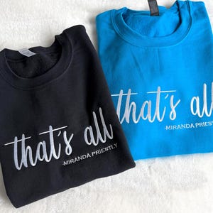May include: Two crewneck sweatshirts, one black and one turquoise blue, are displayed. The phrase "that's all" is printed in white script on both, with "-MIRANDA PRIESTLY" below. The sweatshirts are folded neatly.