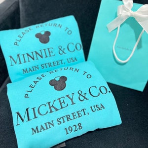 May include: Two turquoise blue tote bags with black text and a black Mickey Mouse silhouette. The text on the bags reads "Please return to Minnie & Co. Main Street, USA" and "Please return to Mickey & Co. Main Street, USA 1928".