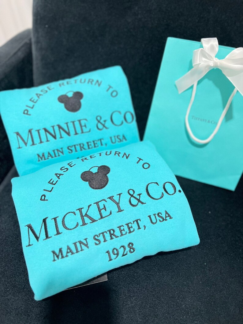 May include: Two turquoise blue tote bags with black text and a black Mickey Mouse silhouette. The text on the bags reads "Please return to Minnie & Co. Main Street, USA" and "Please return to Mickey & Co. Main Street, USA 1928".