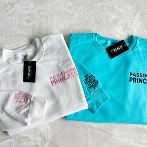 Op de afbeelding: Twee sweatshirts met de tekst "Passenger Princess" erop gedrukt. E&eacute;n sweatshirt is wit met roze tekst en de andere is turquoise met zwarte tekst. Het witte sweatshirt heeft de tekst "He drives while I look pretty" op de mouw gedrukt.