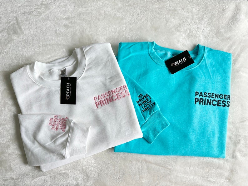 Op de afbeelding: Twee sweatshirts met de tekst "Passenger Princess" erop gedrukt. E&eacute;n sweatshirt is wit met roze tekst en de andere is turquoise met zwarte tekst. Het witte sweatshirt heeft de tekst "He drives while I look pretty" op de mouw gedrukt.