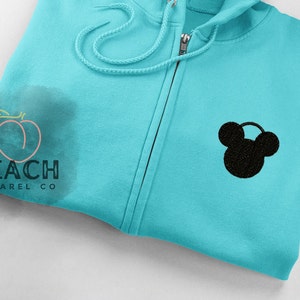 May include: A turquoise blue zippered hoodie with a black Mickey Mouse silhouette embroidered on the front. The hoodie has a drawstring hood and a zipper closure.