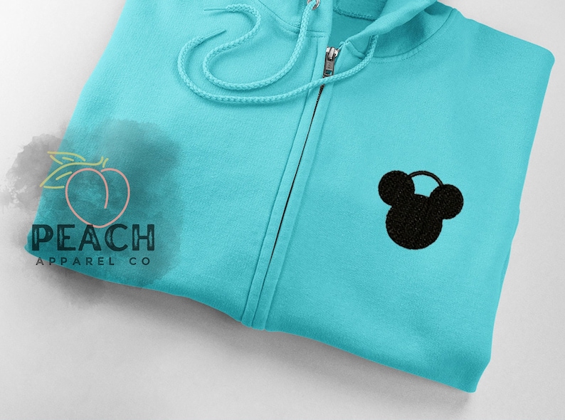 May include: A turquoise blue zippered hoodie with a black Mickey Mouse silhouette embroidered on the front. The hoodie has a drawstring hood and a zipper closure.