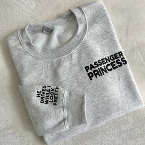 Op de afbeelding: Grijs sweatshirt met de tekst "Passenger Princess" aan de rechterkant en "He drives while I look pretty" op de linkermouw.