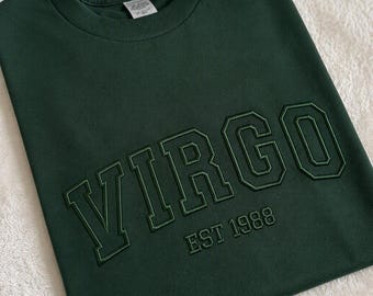 Pode incluir: Uma t-shirt de gola redonda em verde floresta escuro com a palavra VIRGO em letras maiúsculas grandes, a negrito e contornadas no peito, com o texto EST 1988 posicionado logo abaixo num estilo de letra e cor correspondentes.