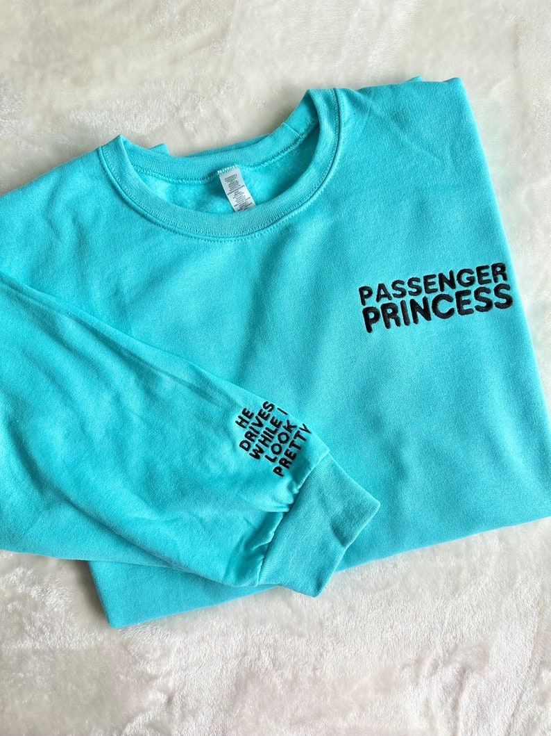 May include: A turquoise blue long-sleeved sweatshirt with the text 'PASSENGER PRINCESS' on the right side of the chest and the text 'HE DRIVES WHILE I LOOK PRETTY' on the left sleeve.