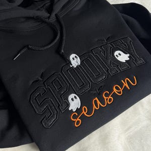May include: A black hooded sweatshirt featuring embroidered text that reads SPOOKY in large black block letters and season in orange cursive script. Three small white ghost figures are positioned around the text, along with small embroidered bats.