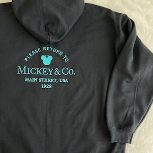 May include: Black hooded sweatshirt with a turquoise blue Mickey Mouse logo and text that reads "Please return to Mickey & Co. Main Street, USA 1928".