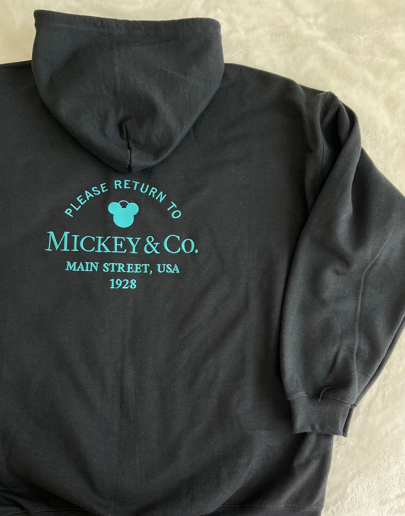May include: Black hooded sweatshirt with a turquoise blue Mickey Mouse logo and text that reads "Please return to Mickey & Co. Main Street, USA 1928".
