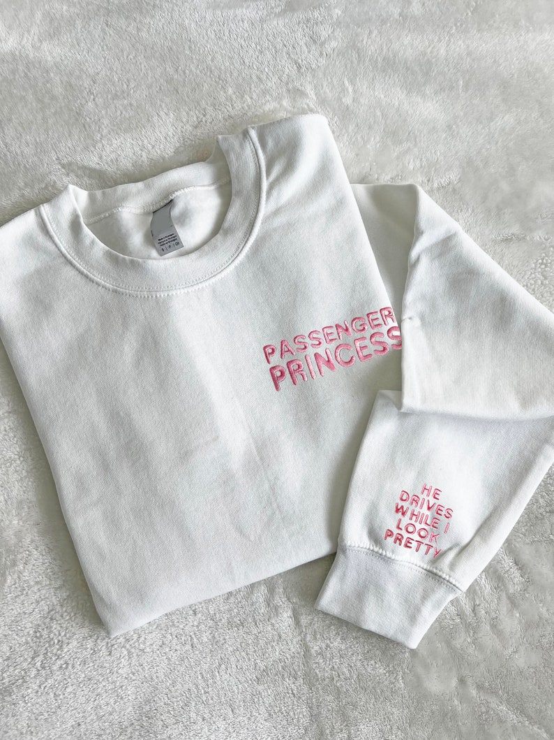 Op de afbeelding: Witte trui met roze geborduurde tekst die "Passenger Princess" op de voorkant en "He drives while I look pretty" op de mouw staat.
